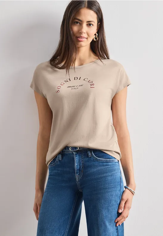 Frau im beigen T-Shirt mit Schriftzug und blauen Jeans vor hellem Hintergrund.