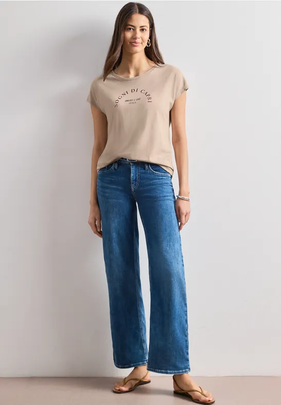 Frau in beigem T-Shirt, weiten blauen Jeans und Sandalen, vor weißem Hintergrund.