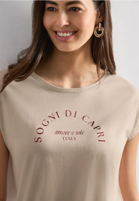 Fröhliche Frau mit hellbeigem T-Shirt mit Aufdruck 'SOGNI DI CAPRI amore e sole ITALY' und goldenen Creolen