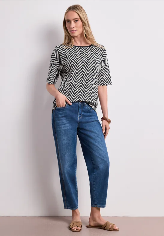 Person trägt schwarz-weißes Zickzack-Shirt, weite blaue Jeans und braune Sandalen vor weißem Hintergrund.