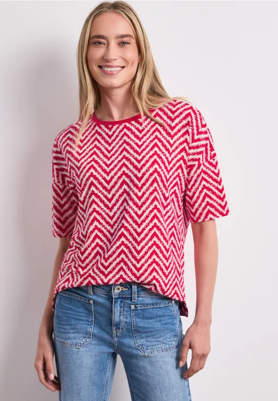 Frau im rot-weißem Zickzack-Shirt und blauen Jeans vor hellem Hintergrund.