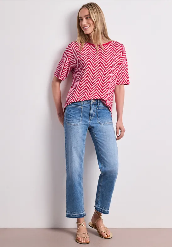 Frau in rotem Zickzack-T-Shirt, blauen Cropped-Jeans und goldenen Sandalen vor neutralem Hintergrund.