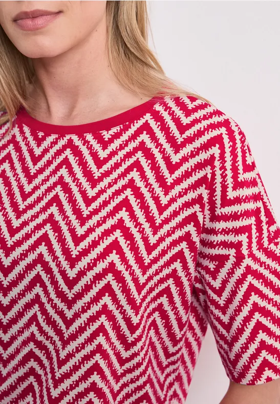 Frau trägt rotes T-Shirt mit weißem Zickzackmuster, halblange Ärmel und Rundhalsausschnitt.