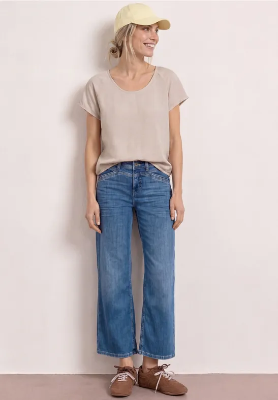 Une personne est debout devant un fond blanc, portant un t-shirt beige, un jean bleu droit et des chaussures marron.