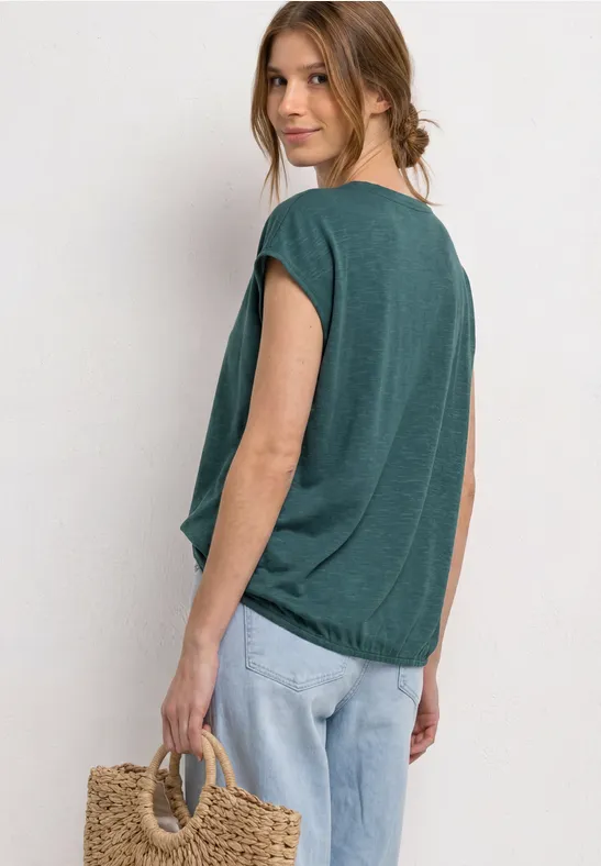 Femme en t-shirt ample vert émeraude et jean, vue de côté avec un sac en paille sur fond clair