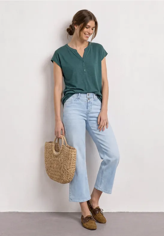 Personne en chemisier vert, jean bleu clair et mocassins bruns, tenant un sac en osier devant un mur blanc