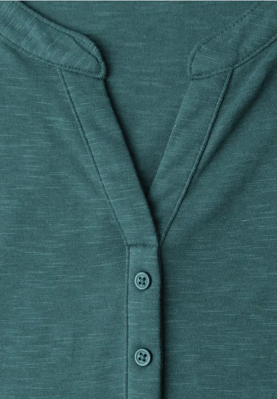Gros plan d’un t-shirt col tunisien bleu-vert à manches longues avec patte de boutonnage et coutures décoratives.