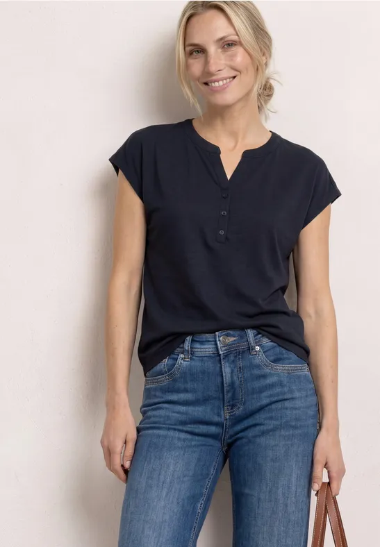 Frau in dunkelblauer Bluse und Jeans steht vor hellem Hintergrund und hält eine Tasche in der Hand.