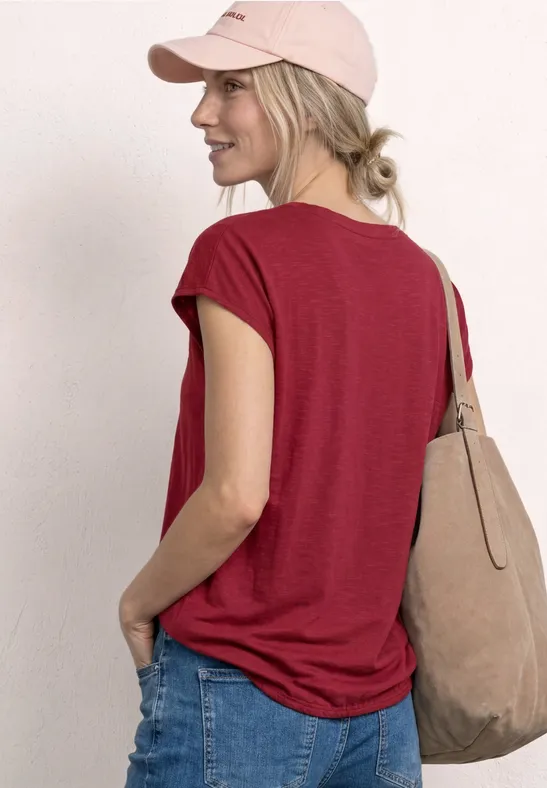 Vrouw van achteren in rood shirt, spijkerbroek, roze pet en beige tas.