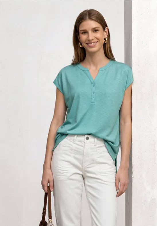 Femme en t-shirt vert menthe à col V et pantalon chino blanc, tenant un sac à main marron.