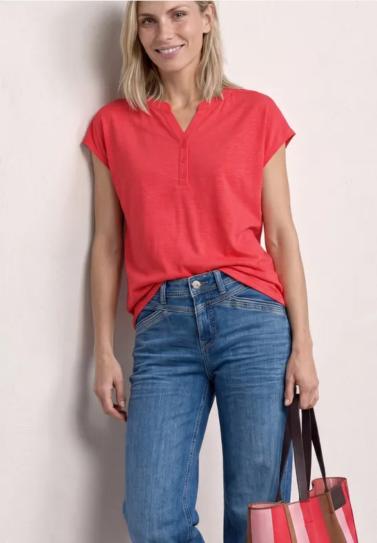 Frau in rotem Top und blauen Jeans mit Handtasche, lächelnd vor neutralem Hintergrund.