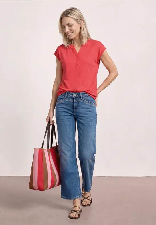 Frau mit rotem Poloshirt, blauen Jeans und gestreifter Tragetasche steht in Sandalen vor hellem Hintergrund.