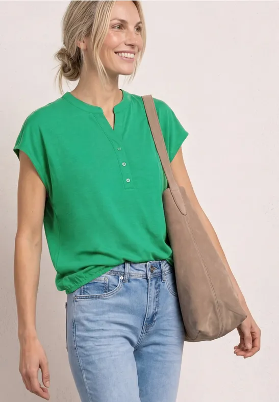 Vrouw in groen T-shirt, blauwe jeans en beige tas bij neutrale achtergrond.