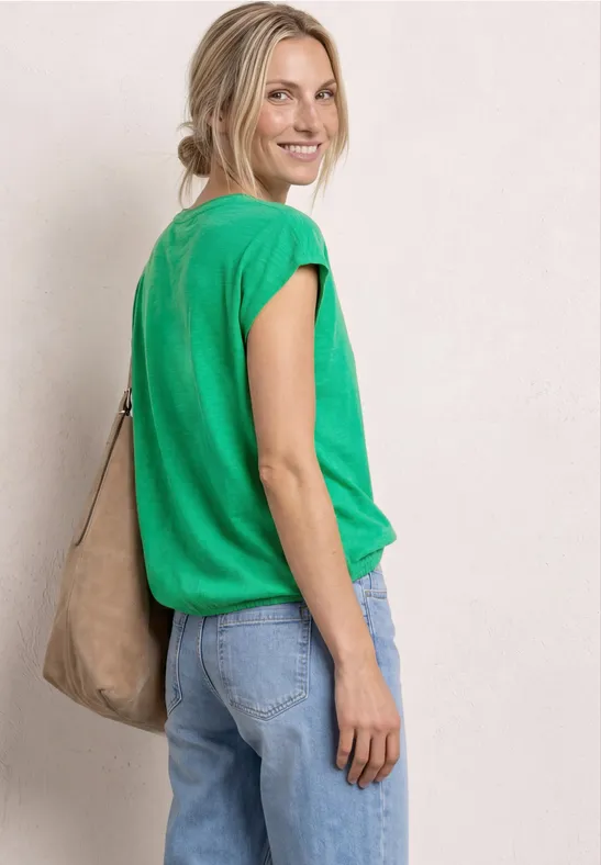 Vrouw in profiel met groen shirt, blauwe spijkerbroek en beige schoudertas voor neutrale achtergrond.