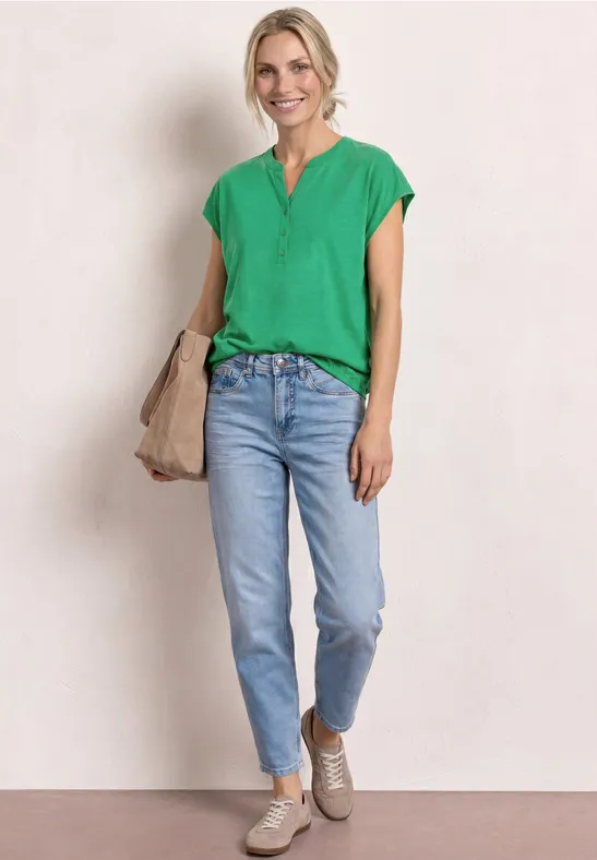 Vrouw in groen shirt en lichtblauwe jeans, met beige tas en schoenen voor lichte achtergrond.