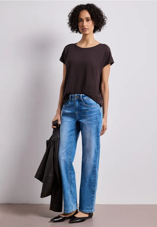 Eine Person steht mit Jeans, dunklem T-Shirt und hält eine schwarze Jacke in der Hand.