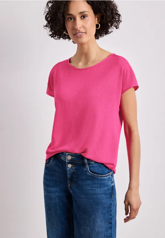 Een vrouw draagt een roze t-shirt en blauwe spijkerbroek tegen een witte achtergrond.