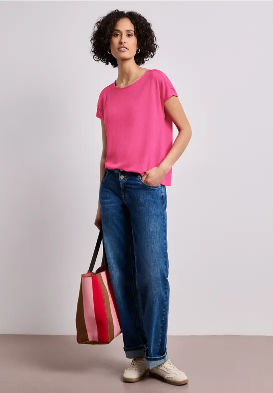 Een persoon draagt een roze T-shirt, brede blauwe spijkerbroek en houdt een gestreepte tas vast.