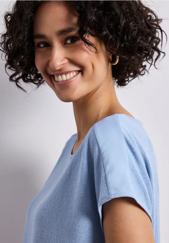 Een persoon met krullend haar draagt een lichtblauw T-shirt met korte mouwen.