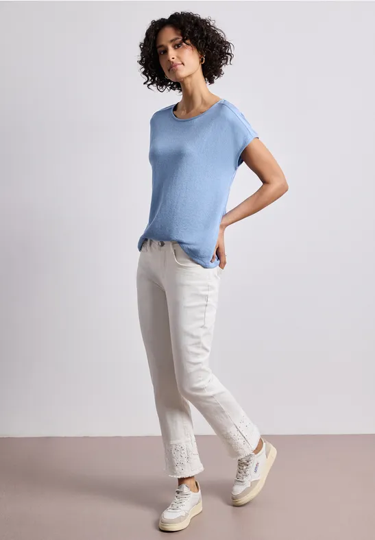 Vrouw voor een neutrale achtergrond, met lichtblauw shirt, witte jeans en witte sneakers.
