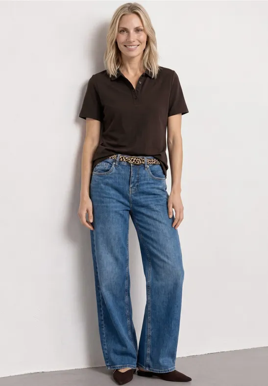 Stehende Person in schwarzem Poloshirt, weitem Jeans, Leoparden-Gürtel und braunen Schuhen vor weißem Hintergrund.