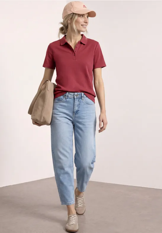 Vrouw in rood poloshirt, lichtblauwe jeans, beige schoenen en tas tegen neutrale achtergrond.