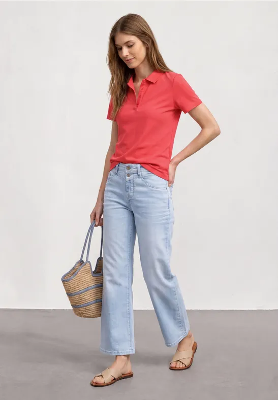 Frau in rotem Poloshirt, hellblauen Jeans und Sandalen, hält eine geflochtene Tasche.