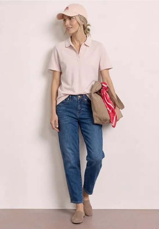 Vrouw staat voor een lichte muur, draagt licht poloshirt, blauwe jeans en lage schoenen, houdt een rode tas losjes vast.