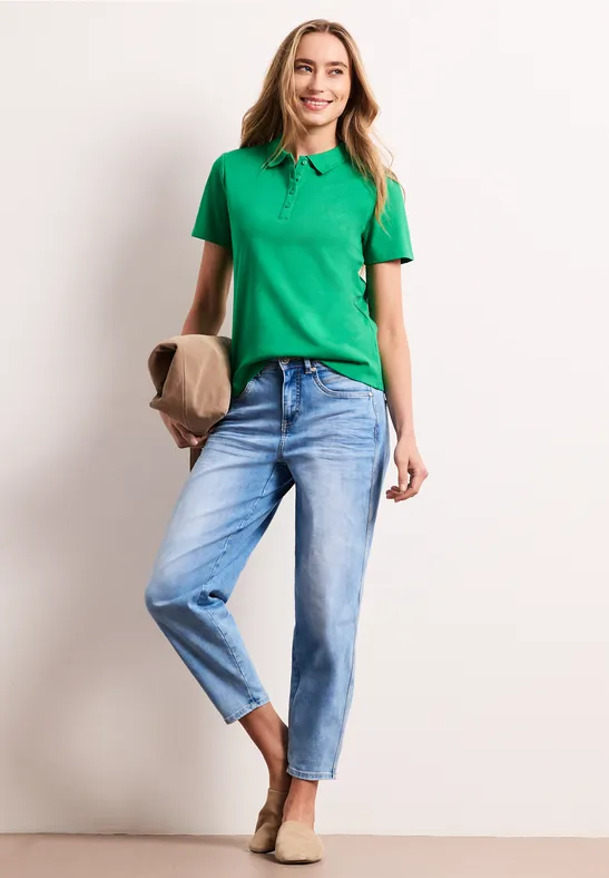 Vrouw voor witte muur in groen poloshirt, blauwe jeans en comfortabele schoenen, houdt clutch vast.