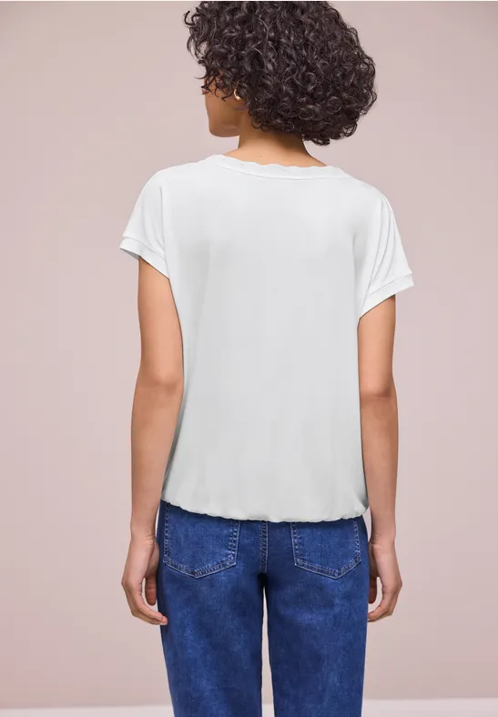 Frau mit kurzen Locken von hinten in weißem T-Shirt und blauen Jeans vor neutralem Hintergrund.