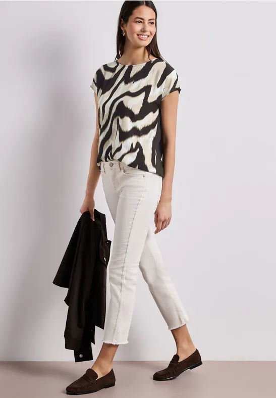 Vrouw in zebra-top, witte broek en loafers, met een zwarte jas in de hand.