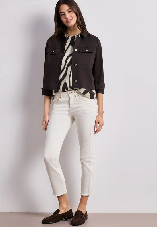 Vrouw in witte jeans, zwarte korte jas, zebra-print blouse en bruine loafers voor een lichte achtergrond.