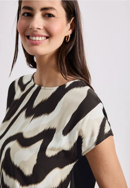 Een persoon draagt een shirt met korte mouwen en abstract zwart-crèmemotief tegen een effen achtergrond.