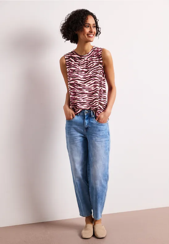 Persoon in rood-wit gestreepte mouwloze top, blauwe jeans en beige schoenen, ontspannen staand met handen in de zakken.