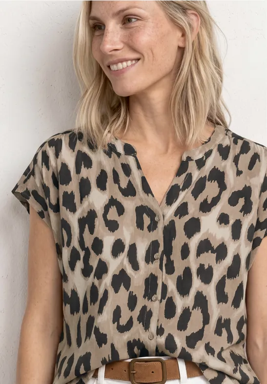 Frau mit beige-schwarz gemusterter Bluse im Leoparden-Print, kurzem Ärmel und braunem Gürtel, lächelnd.