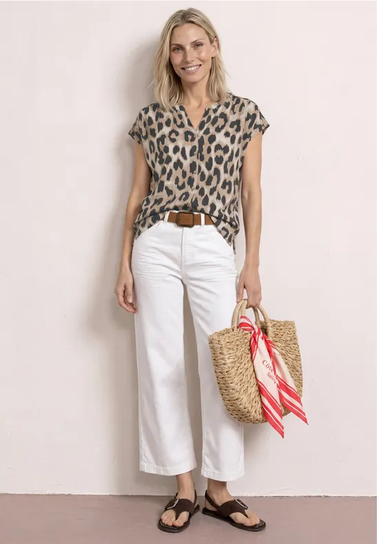 Frau trägt Leopardenmuster-Bluse, weiße Culotte, flache Sandalen und Strohtasche mit rotem Tuch.