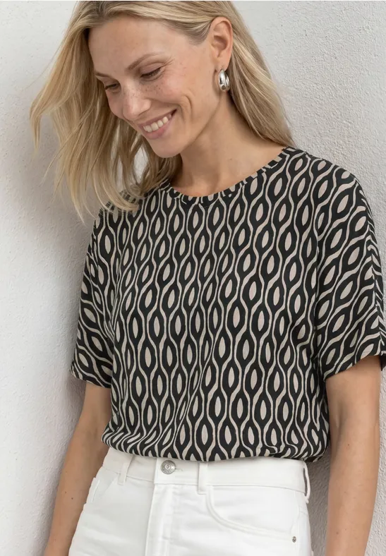Frau trägt gemustertes, schwarz-beige Oberteil und weiße Hose.