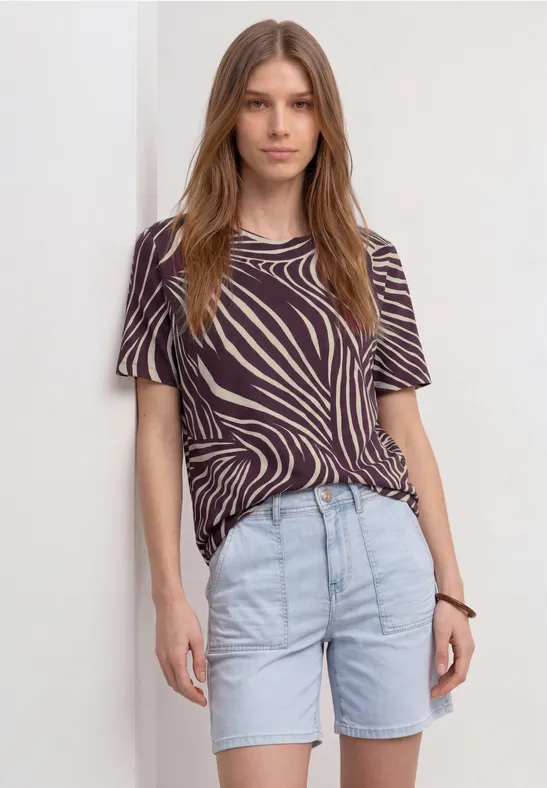 Person trägt zebramuster-T-Shirt, helle Jeans-Shorts und Armband, steht vor weißem Hintergrund.
