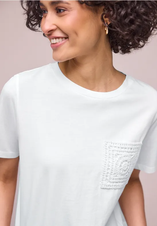 Vrouw met krullend haar en onscherp gezicht draagt een wit T-shirt met gehaakt zakje.