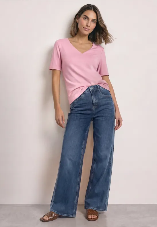 Frau in rosa V-Shirt und weiten Jeans steht vor neutralem Hintergrund.