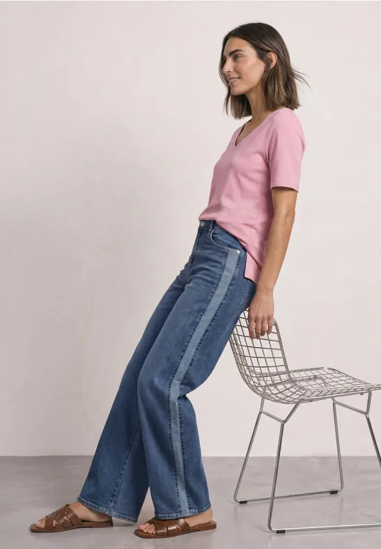 Frau in rosa T-Shirt und zweifarbigen Jeans lehnt an modernem Drahtstuhl vor weißem Hintergrund.