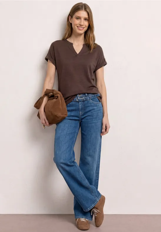 Femme posant devant un mur clair, t-shirt marron, jean bleu, chaussures marron et sac en daim.