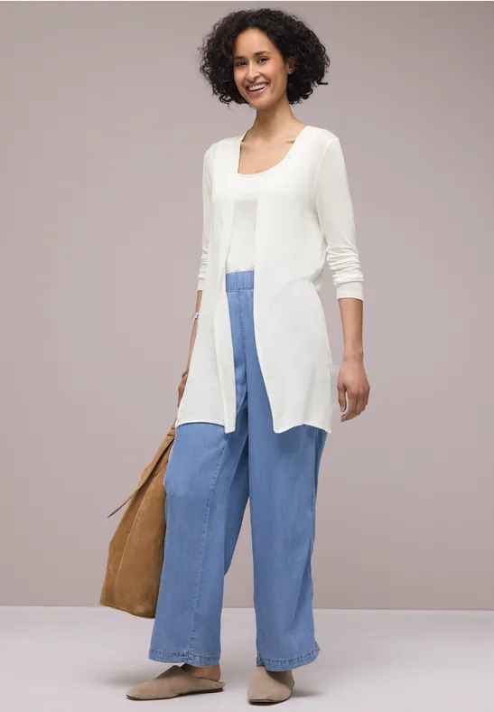 Een persoon draagt een lange witte cardigan, wijde blauwe broek, beige espadrilles en een bruine tas.