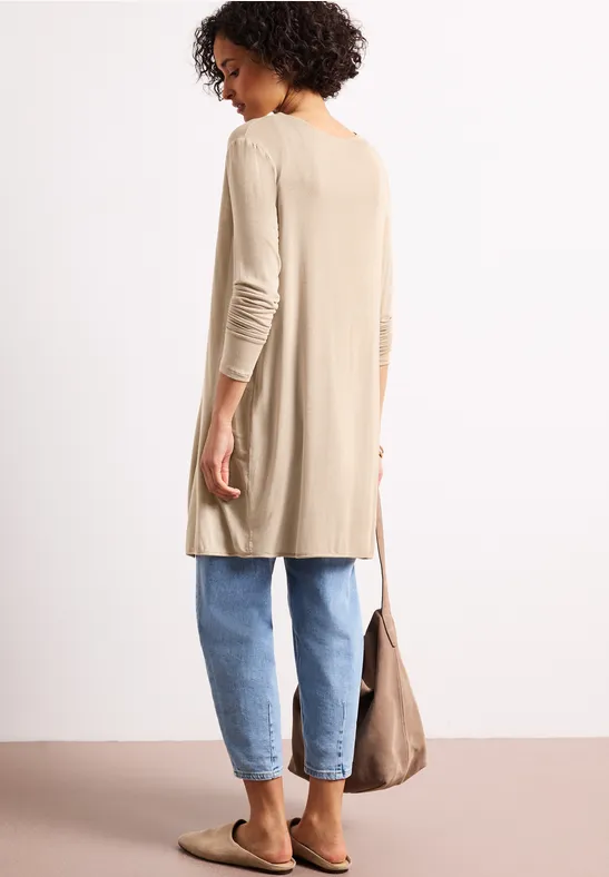 Achteraanzicht van een vrouw in een beige lange cardigan, blauwe jeans en instappers, met een beige tas