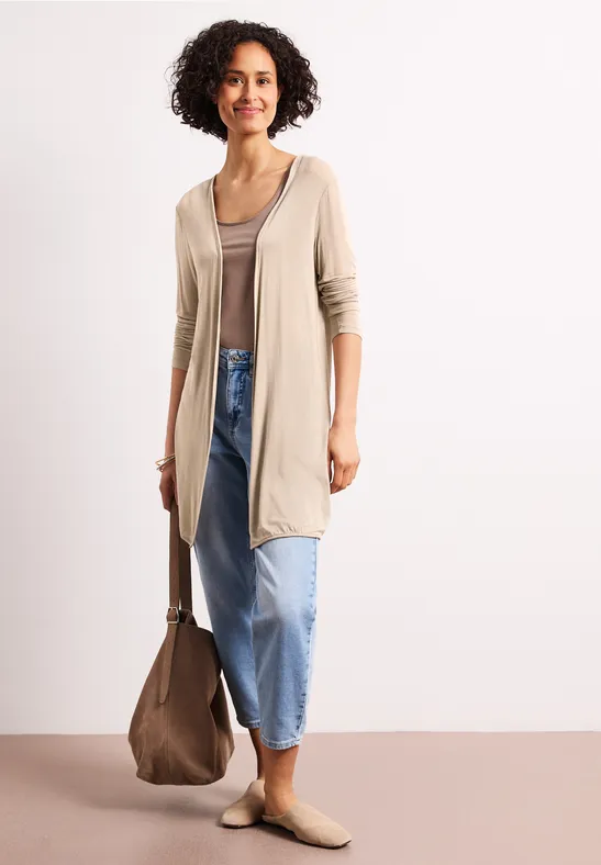 Vrouw in beige vestje en top, blauwe jeans, bruine instappers en passende tas voor neutrale achtergrond.