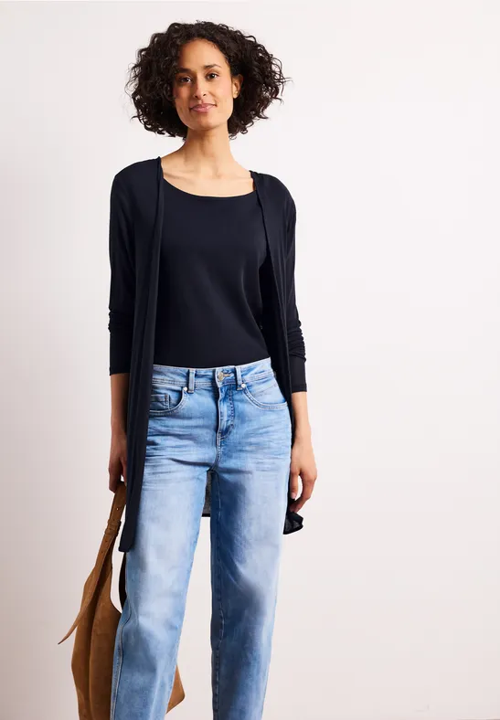 Een vrouw met krullend haar draagt een zwarte top, een vest, een lichtblauwe spijkerbroek en een bruine tas tegen een witte achtergrond.