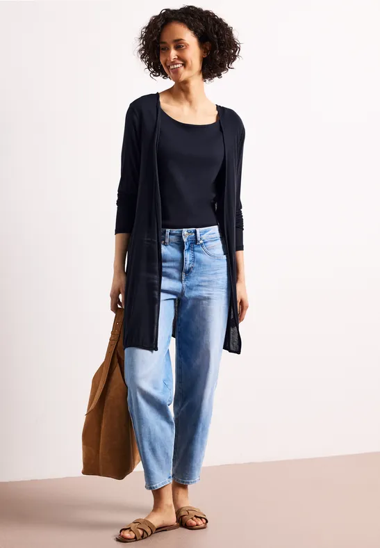 Vrouw in zwarte top en vest, losse blauwe jeans, sandalen en grote tas.