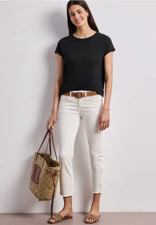 Frau in schwarzem Shirt, weißen Hosen, flachen Sandalen und Flecht-Tasche vor weißer Wand.