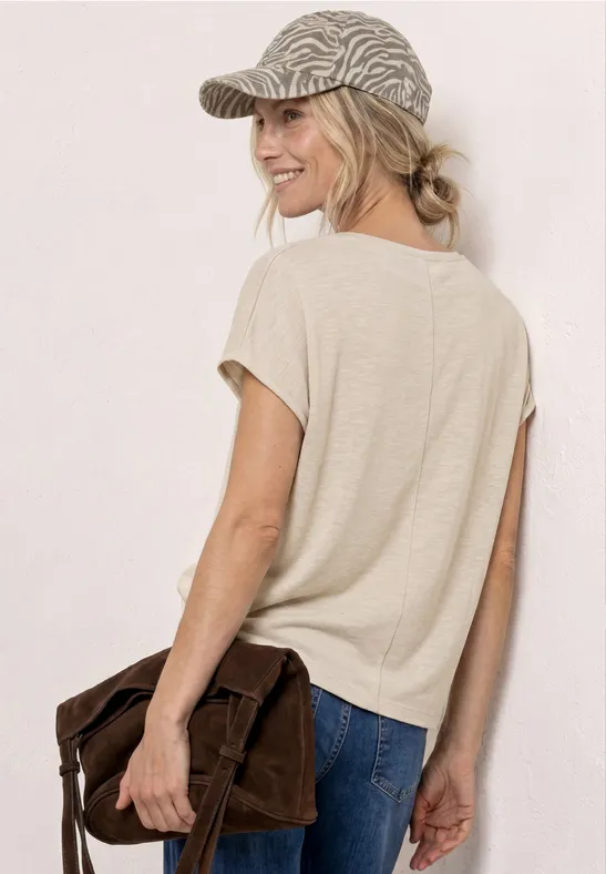Vrouw van achteren in een casual outfit: beige T-shirt, jeans, pet en bruine schoudertas.