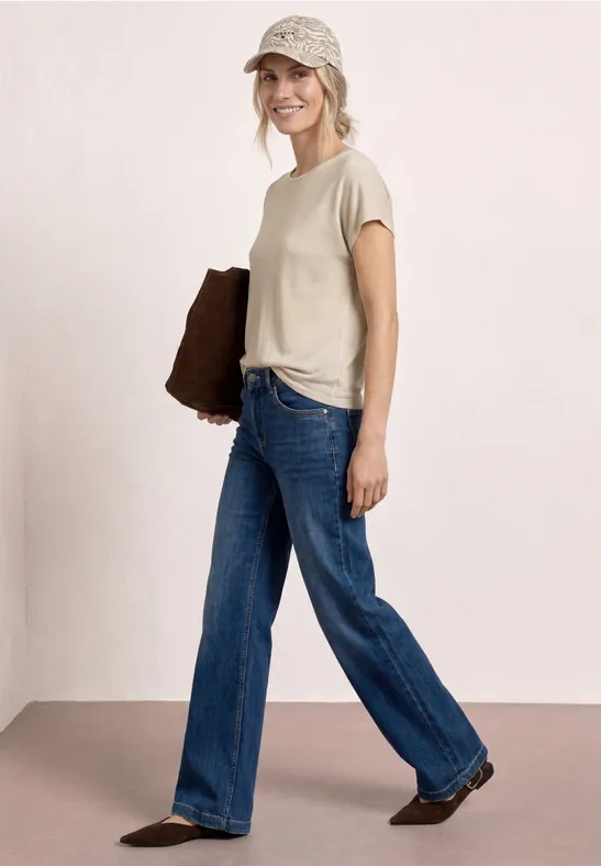 Vrouw in beige T-shirt, wijde jeans, bruine tas en hoofddoek in een nonchalante houding.